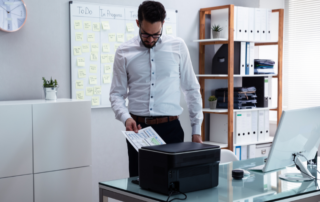 Young professional man works in an office at a desk grabbing printouts from his office equipment.