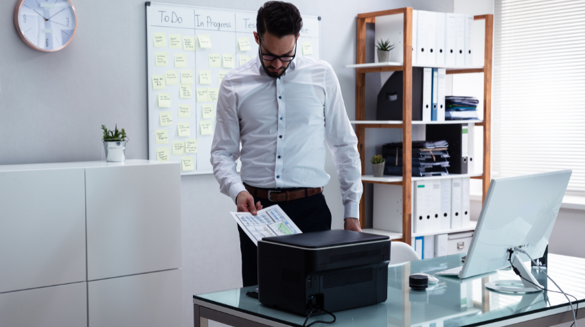 Young professional man works in an office at a desk grabbing printouts from his office equipment.