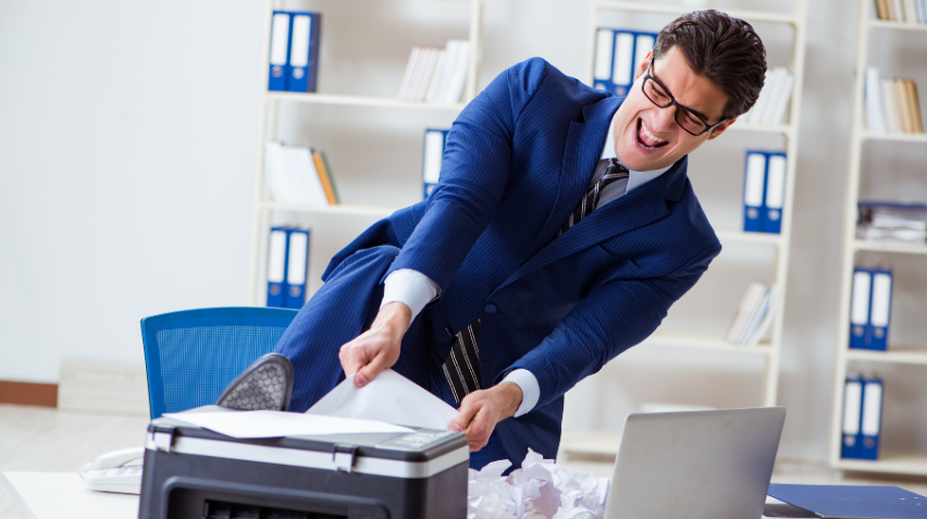 An office worker frowning while looking at a printer, trying to resolve printer issues. The printer displays an error message indicating a paper jam, with scattered sheets of paper lying around.