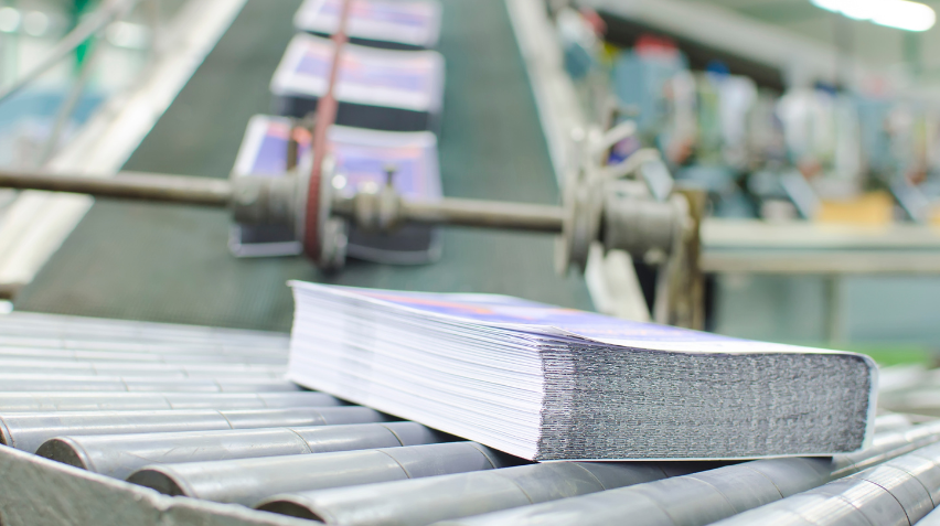 Stack of print books moving along a conveyor belt in a printing facility, with blurred machinery in the background. The glossy print books are neatly bound, ready for distribution.