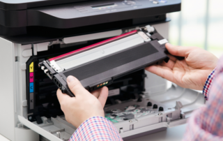 Person replacing a printer toner cartridge in a color laser printer, with yellow, magenta, cyan, and black toner slots visible.