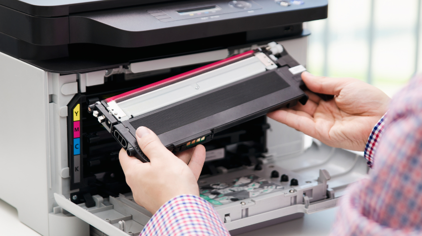 Person replacing a printer toner cartridge in a color laser printer, with yellow, magenta, cyan, and black toner slots visible.